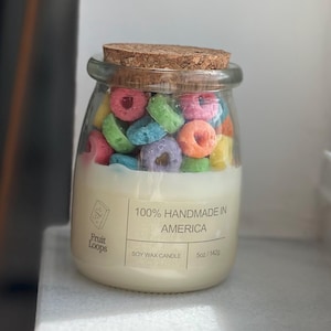 May include: A clear glass jar candle filled with colorful cereal and soy wax. The candle is labeled "100% Handmade in America" and "Fruit Loops" with a 5oz/142g weight. The jar has a cork lid.