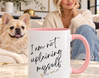 May include: A white ceramic mug with a light pink interior and handle. The front features black cursive text that reads I am not explaining myself, followed by smaller text that reads now f*ck off and a small gold heart outline.