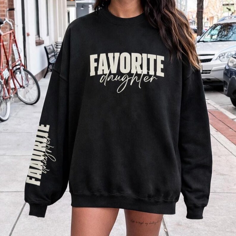 May include: Black sweatshirt featuring the text "FAVORITE daughter" in white. The words are in block letters across the chest and vertically down the left sleeve. A casual, comfortable style.