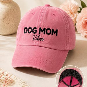 May include: A rose-colored baseball cap featuring the embroidered black text DOG MOM Vibes on the front. The cap has a curved brim and a button on top. An inset shows the interior of the cap, which is lined with a black fabric featuring white polka dots.