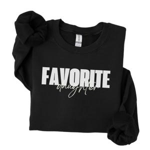 May include: Black sweatshirt with the text "FAVORITE daughter" in white block letters and cursive. The sweatshirt has long sleeves and a crew neck. The sleeves are rolled up. The Gildan brand tag is visible.