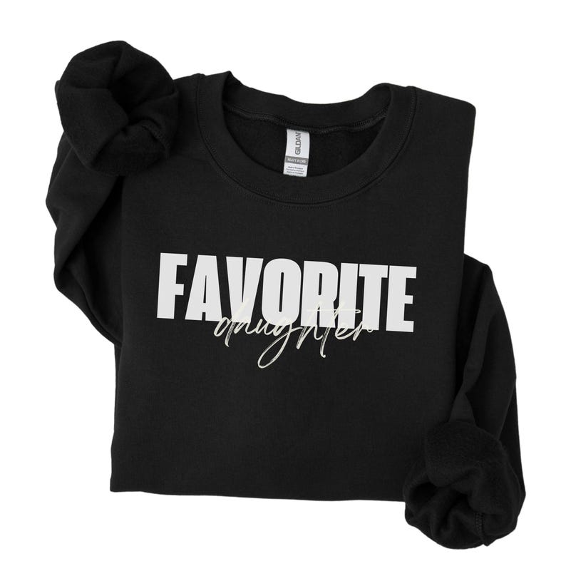 May include: Black sweatshirt with the text "FAVORITE daughter" in white block letters and cursive. The sweatshirt has long sleeves and a crew neck. The sleeves are rolled up. The Gildan brand tag is visible.