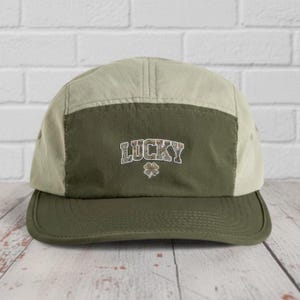 May include: A green and beige five-panel cap with a flat peak. The front panel is olive green with the word "LUCKY" in a textured font and a small shamrock. The cap is made of a lightweight material.