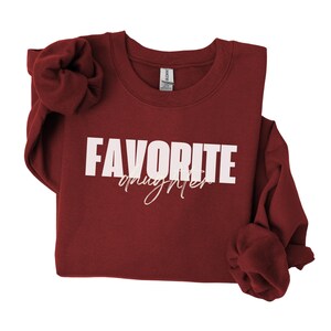 May include: A maroon sweatshirt featuring the text "FAVORITE" in bold white capital letters and "daughter" in a cursive font. The long-sleeved top is made of a soft material.