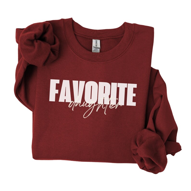 May include: A maroon sweatshirt featuring the text "FAVORITE" in bold white capital letters and "daughter" in a cursive font. The long-sleeved top is made of a soft material.
