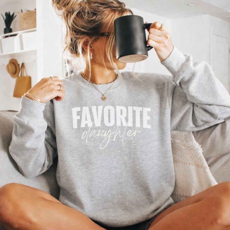 May include: A light grey sweatshirt with the words "FAVORITE daughter" printed in white. The person is holding a black mug. The sweatshirt has long sleeves and a crew neck.