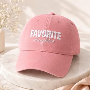 K&ouml;nnte beinhalten: Rosa Baseballkappe mit dem gestickten Text "FAVORITE daughter" in Wei&szlig;. Die Kappe hat einen gebogenen Schirm und eine abgerundete Oberseite. Der Hut ist aus einem weichen Stoff gefertigt und ein l&auml;ssiges Accessoire.