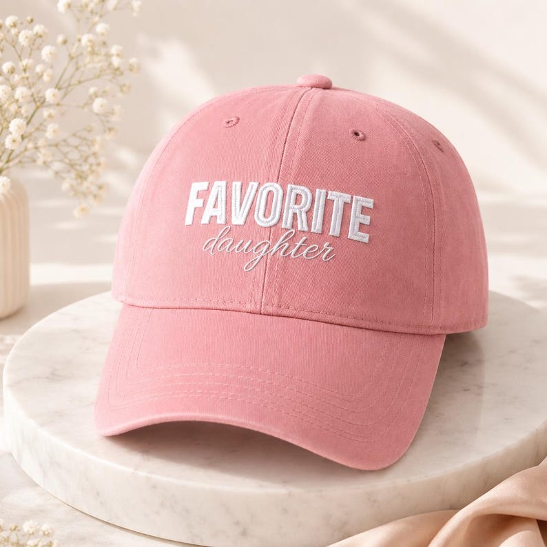 K&ouml;nnte beinhalten: Rosa Baseballkappe mit dem gestickten Text "FAVORITE daughter" in Wei&szlig;. Die Kappe hat einen gebogenen Schirm und eine abgerundete Oberseite. Der Hut ist aus einem weichen Stoff gefertigt und ein l&auml;ssiges Accessoire.