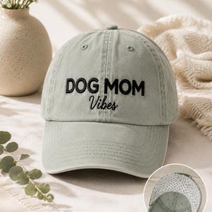 May include: A sage green baseball cap featuring the embroidered black text "DOG MOM Vibes" on the front. The cap has a curved brim and a small circular inset showing the interior lining, which is white with a black polka dot pattern.