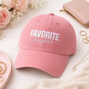 K&ouml;nnte beinhalten: Staubrosa Baseballkappe mit dem gestickten Text "FAVORITE daughter" in Wei&szlig;. Die Kappe ist aus einem weichen Stoff gefertigt und hat einen gebogenen Schirm. Accessoires wie eine Geldb&ouml;rse, Ringe und eine Halskette sind im Hintergrund.