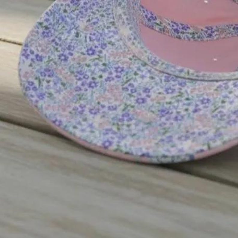 K&ouml;nnte beinhalten: Nahaufnahme einer Baseballkappe mit floralem Muster. Die Kappe hat ein rosa Innenfutter und einen rosa Rand, und die Au&szlig;enseite ist mit kleinen lila und rosa Blumen auf wei&szlig;em Grund gemustert. Die Kappe liegt auf einer Holzoberfl&auml;che.