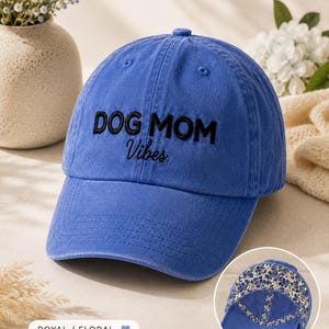 May include: A royal blue baseball cap featuring the embroidered black text DOG MOM Vibes on the front. The interior lining of the cap displays a multi-colored floral pattern with blue, yellow, and white flowers on a cream background.