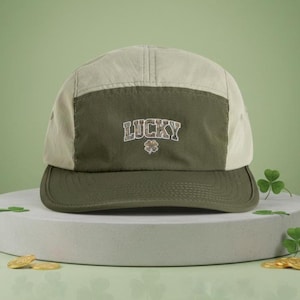 May include: A green and beige baseball cap with the word "LUCKY" in a textured font and a shamrock graphic. The cap has a curved brim and a five-panel design. The background is a light green.