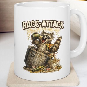 May include: White ceramic mug featuring a raccoon in a trash can, holding a can and pizza. The mug has the text "RACC-ATTACK" in a brown font. The raccoon is surrounded by trash and food items.