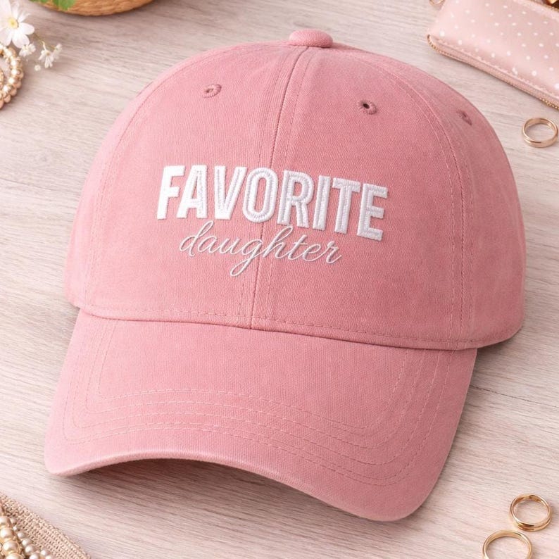 K&ouml;nnte beinhalten: Eine rosa Baseballkappe mit den Worten "FAVORITE daughter" in Wei&szlig; gestickt. Die Kappe hat einen gebogenen Schirm und einen verstellbaren Riemen. Der Hut ist aus einem weichen Stoff gefertigt.