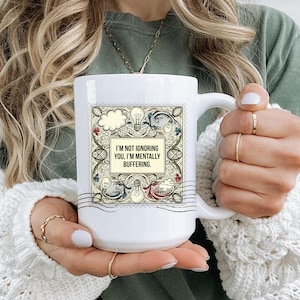 Puede incluir: Taza de cerámica blanca con asa, con el texto "I'M NOT IGNORING YOU, I'M MENTALLY BUFFERING." La taza tiene un borde decorativo con ilustraciones de bombillas y nubes. La taza está sujeta por una persona.