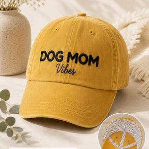 May include: A sunset yellow baseball cap featuring the embroidered black text DOG MOM Vibes on the front. The interior lining of the cap is white with black polka dots, as shown in the circular inset image at the bottom right corner.