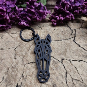 May include: Black wooden horse head keyring with a geometric design. The keyring is on a wooden surface with purple flowers in the background.