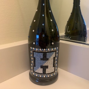 May include: A black glass bottle with a blue and silver rhinestone design. The bottle has a square-shaped design on the front with the letter 