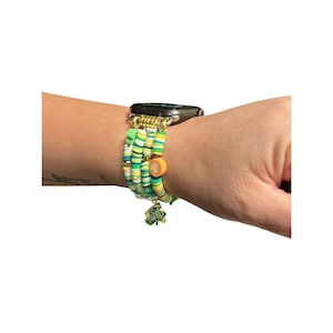May include: A green, white, and yellow beaded Apple Watch band. The band features a charm of a green shamrock. The band is attached to a black Apple Watch. The band is on a wrist.