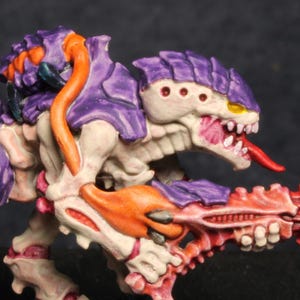May include: A purple and white painted miniature figurine of a creature with a long, thin tongue and a large, orange gun. The creature has a long, thin tail and is standing on a black base.