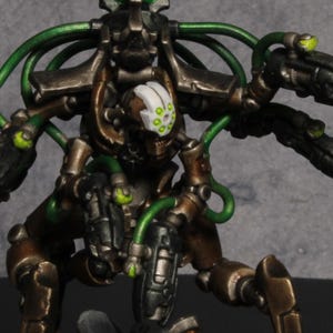 May include: A miniature figurine of a robotic warrior with a brown and black metallic finish. The figure has green accents, including a glowing head and connecting tubes. It stands on a circular base with a textured, brown surface.
