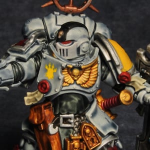 Può includere: Figura in miniatura dettagliata di un marine spaziale in armatura grigia con accenti gialli. La figura tiene in mano una pistola e uno scudo. La miniatura ha un'aureola e si trova su una base marrone. La figura è dipinta con dettagli intricati.