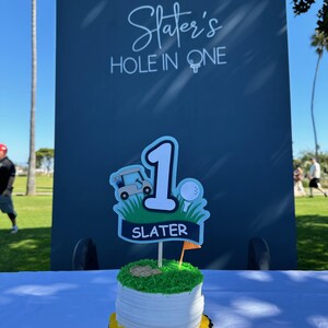 May include: A round cake with white frosting and yellow decorative piping at the base, topped with a golf-themed cake topper featuring a large number one, a golf cart, a golf ball, and a banner with the name SLATER printed in white text.
