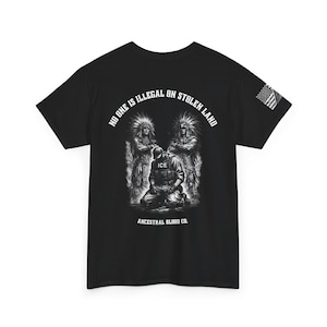 Puede incluir: Camiseta negra con un diseño gráfico que presenta dos figuras nativas americanas que flanquean una figura arrodillada con el texto "ICE" en su chaleco. El texto "NO ONE IS ILLEGAL ON STOLEN LAND" se arquea sobre las figuras, con las palabras "ANCESTRAL BLOOD CO." debajo.
