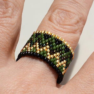 Puede incluir: Un anillo ancho de cuentas con un diseño de camuflaje, que incorpora cuentas verdes, negras y doradas. El anillo, que se lleva en un dedo, muestra un patrón detallado y texturizado. El diseño del anillo es intrincado.