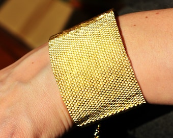 May include: A gold-colored bracelet made of small, tightly woven metal links. The bracelet is worn on a person's wrist.