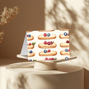 May include: A white card with a repeating pattern of eclairs topped with raspberries and blueberries. The eclairs are filled with white cream and have a light brown crust.