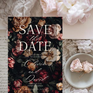 May include: Save the Date wedding invitation with a dark floral background featuring burgundy, pink, and yellow roses. The text reads "Save the Date" in white lettering, followed by "To Celebrate the Wedding of Olivia and David" in cursive script. The date is listed as "Fourth of June Twenty Seventeen".