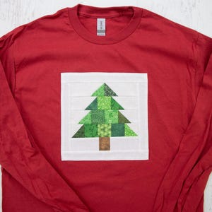 May include: Red long-sleeved t-shirt with a white square featuring a green patchwork quilt tree design.