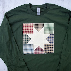 May include: A dark green long-sleeved t-shirt with a patchwork quilt design featuring a white star in the center. The quilt is made of various plaid fabrics in shades of red, blue, green, and white.