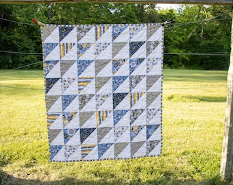 May include: A handmade quilt with a geometric pattern of triangles in white, blue, gray, and yellow fabrics. The quilt is hanging outside on a clothesline, with a green grassy background and trees.