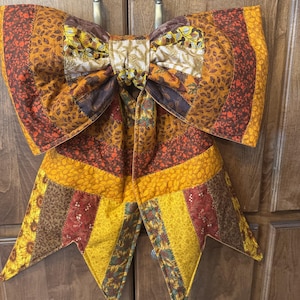 May include: A large, quilted fabric bow featuring a patchwork design in autumn colors including mustard yellow, burnt orange, and deep brown. The fabric displays various patterns of leaves, sunflowers, and floral motifs with stitched seams.