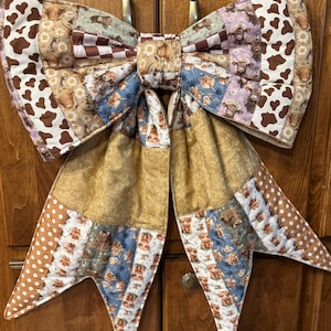 May include: A large fabric bow featuring a patchwork design with various patterns including brown and white cow spots, highland cattle illustrations, daisies, brown and white checkered squares, and tan polka dots, all stitched together in a quilted style.