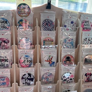 May include: A wooden display rack filled with car coasters. The coasters feature various designs, including text, patterns, and cartoon characters. The coasters are packaged in clear plastic and labeled "Car Coasters".