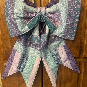 May include: A large decorative bow constructed from quilted fabric patches in shades of purple, teal, and lavender. The bow features a variety of floral and paisley patterns, with two long, pointed tails hanging down from the central knot.