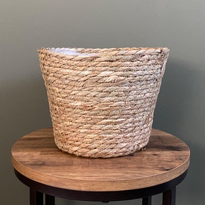 May include: A light brown woven basket with a cylindrical shape, wider at the top. It rests on a round wooden table with a dark metal base. The background is a muted green. The basket is made of natural fibres.