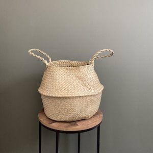 May include: A natural woven basket with two handles, sitting on a wooden stand. The basket is light brown and has a round shape.