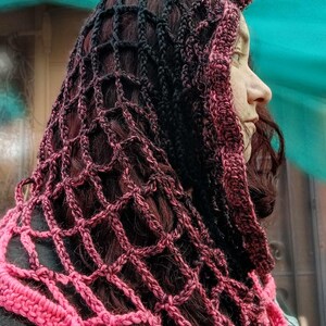 May include: A pink and black crocheted hooded scarf with a delicate openwork pattern. The scarf is made with a gradient yarn that transitions from black to pink.