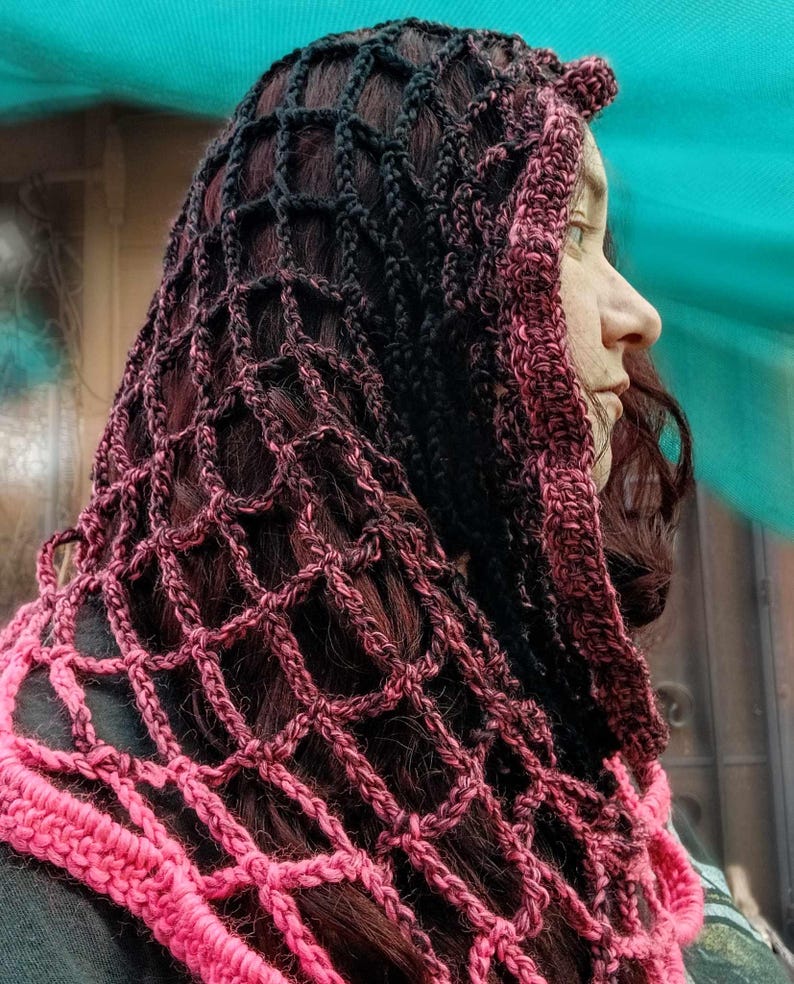 May include: A pink and black crocheted hooded scarf with a delicate openwork pattern. The scarf is made with a gradient yarn that transitions from black to pink.