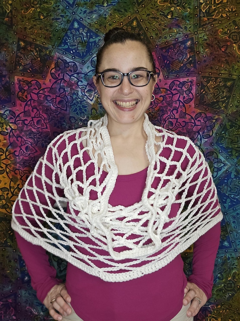 May include: A white crochet shawl with a net-like design. The shawl is draped around the shoulders of a person wearing a pink shirt.