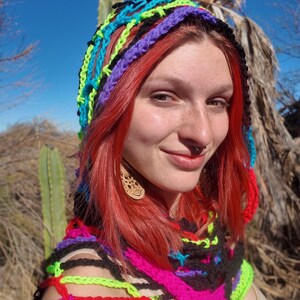 May include: A woman wearing a colorful crocheted hooded scarf with black, pink, blue, green, and purple yarn. The scarf is open in the front and has a loose, flowing design.