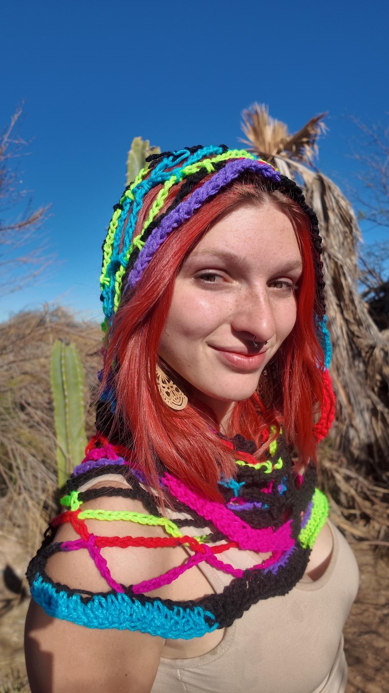 May include: A woman wearing a colorful crocheted hooded scarf with black, pink, blue, green, and purple yarn. The scarf is open in the front and has a loose, flowing design.