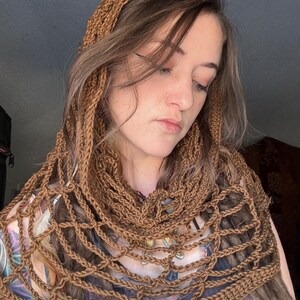 May include: A brown crocheted cowl with a delicate openwork design. The cowl is worn by a person with long brown hair.