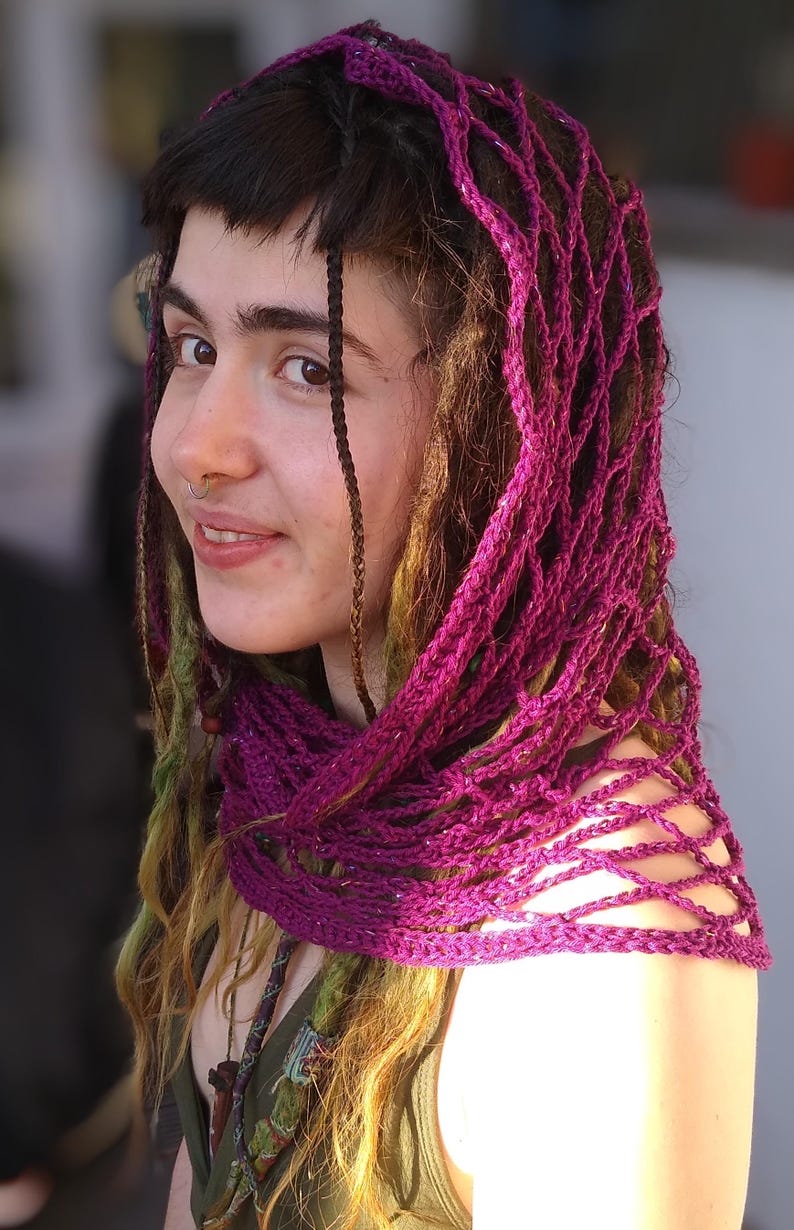 May include: A woman with long, brown hair with purple and green streaks wears a purple crocheted shawl over her head and shoulders. The shawl is a loose, open weave with a delicate pattern.