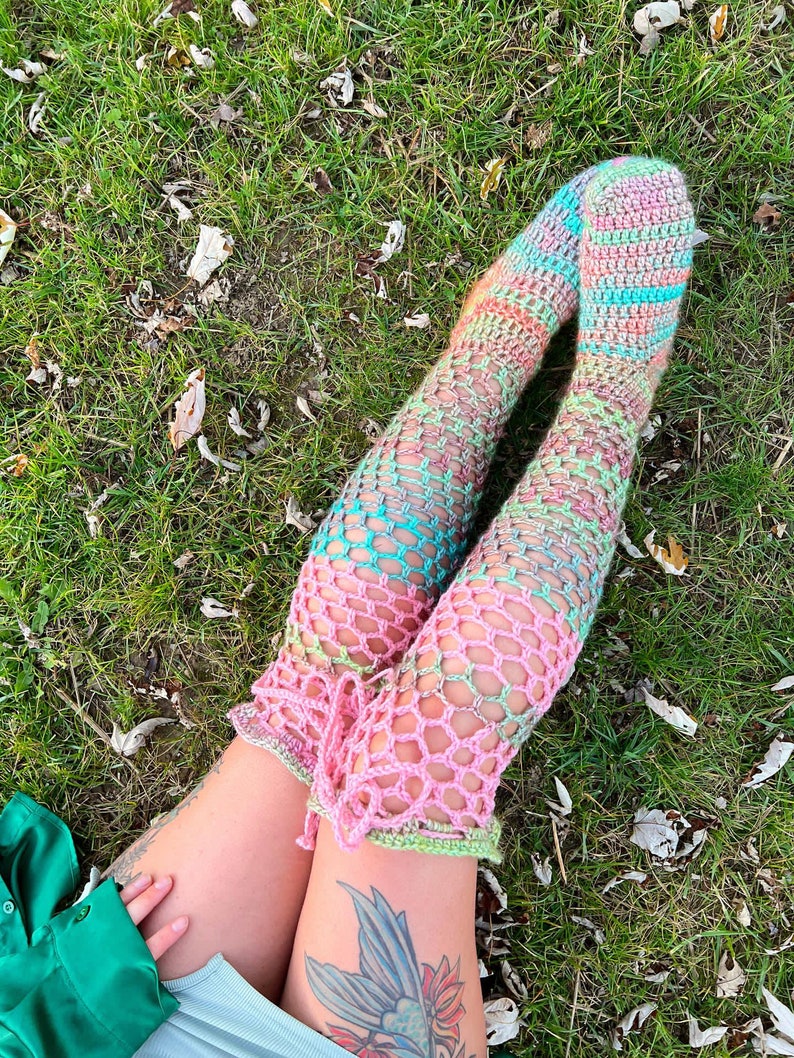 Op de afbeelding: Een paar kleurrijke gehaakte beenwarmers met een kanten patroon. De beenwarmers zijn roze, groen en blauw.